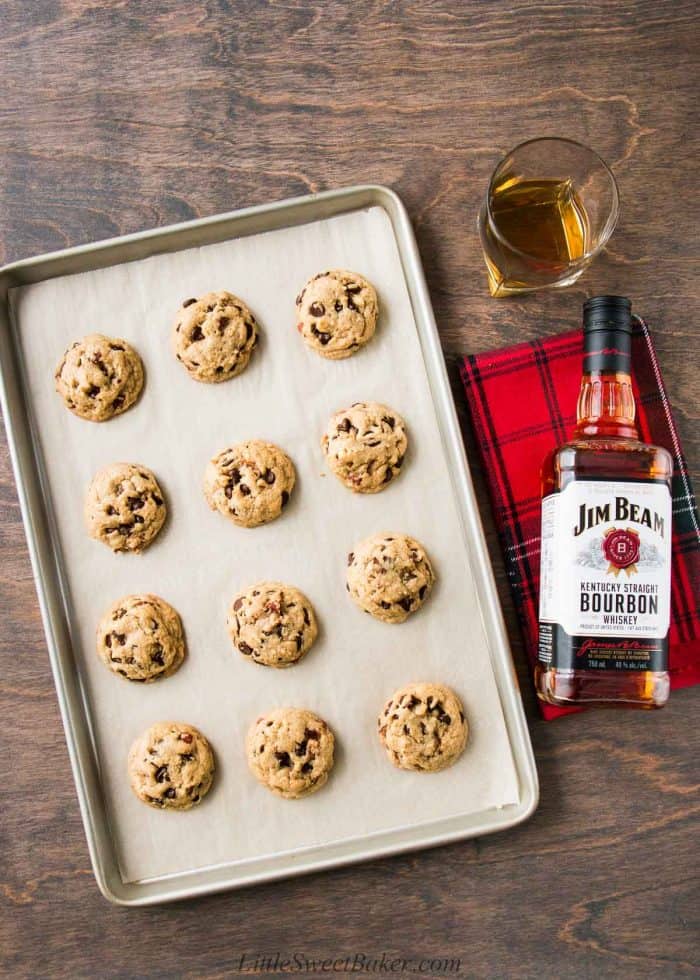 A tray of bourbon chocolate chip cookies with a bottle of bourbon on a wooden table.