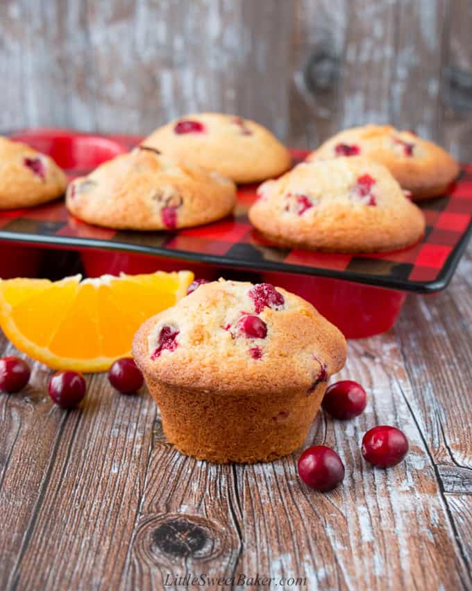 A cranberry orange muffin surrounded by cranberries and an orange wedge on a wooden surface.