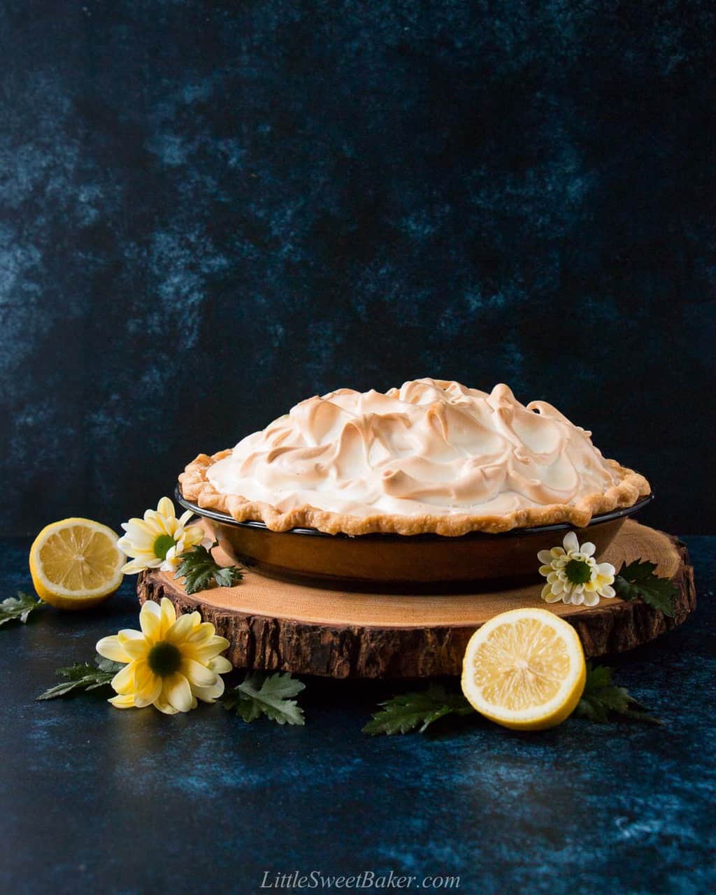 A lemon meringue pie on a wooden board surrounded by flowers and lemons.