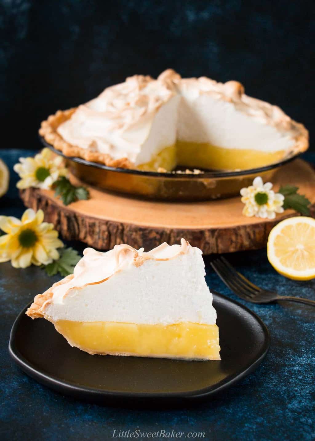 A slice of mile-high lemon meringue pie on a black plate with the rest of the pie behind it.