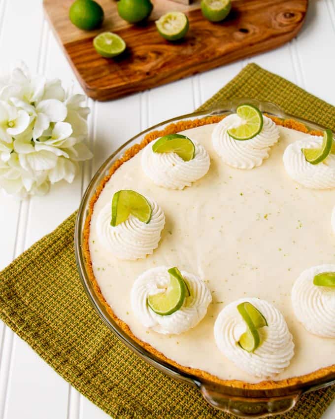 A key lime pie on a green dish towel with white flowers and a cutting board with key limes in the background.