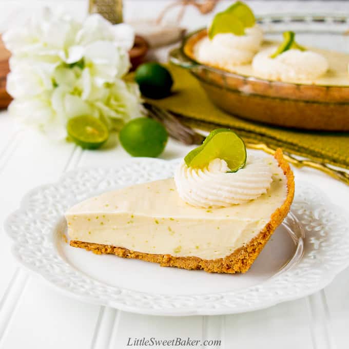 A slice of key lime pie on a white plate with the rest of the pie, white flower and key limes in the background.