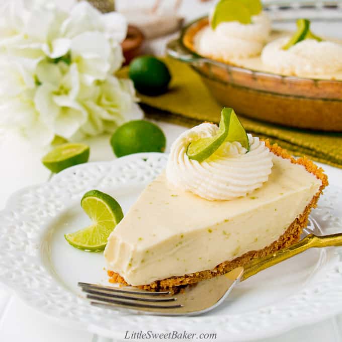 A slice of key lime pie with a bite missing. The rest of the pie, white flowers and key limes in the background.
