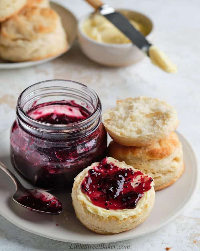 Two biscuits on a plate with a jar of jam. One biscuit with clotted cream and jam on it.