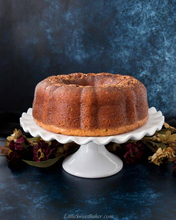 A whole Bacardi rum cake on a white cake stand with flowers and a dark blue background.