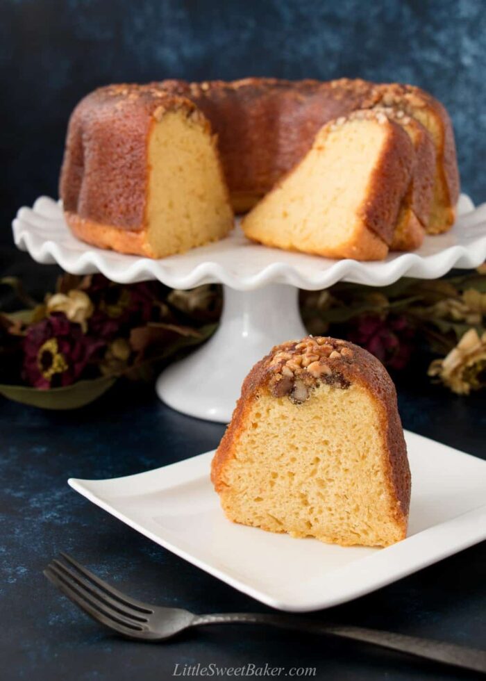 A slice of rum cake on a curved white plate with the rest of the cake on a cake stand behind it.