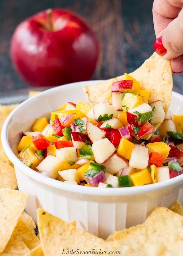 Apple mango salsa in a white bowl surrounded by tortilla chips with a hand dipping a chip.