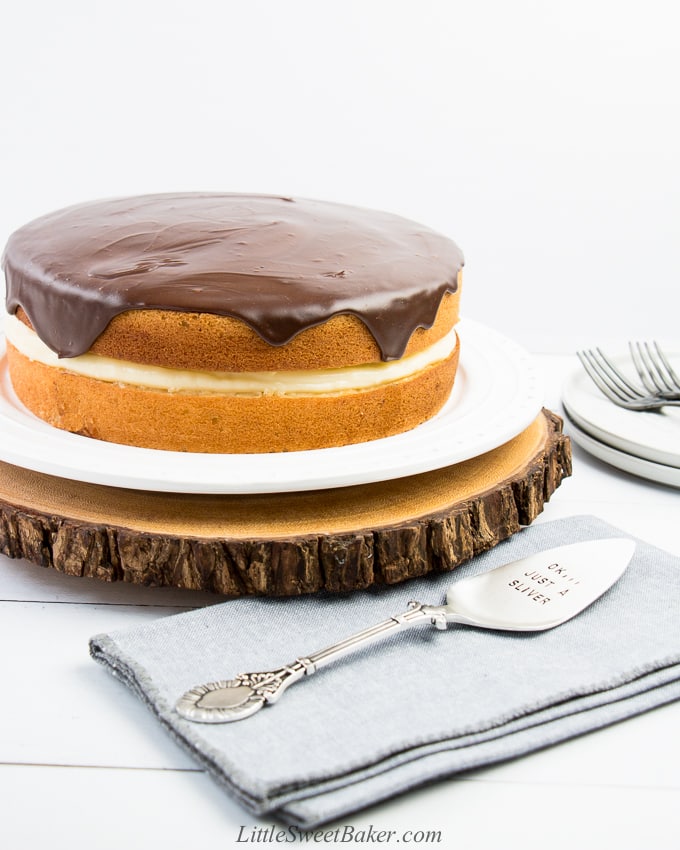 Boston cream pie on a white plate and wooden board with grey napkin and silver cake server.