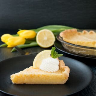A slice of lemon pie on a black plate with whipped cream, lemon wedge and yellow tulips in the background.