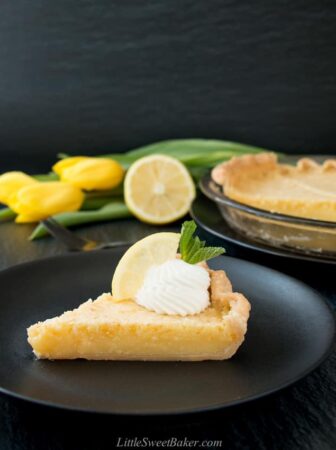 A slice of lemon pie on a black plate with whipped cream, lemon wedge and yellow tulips in the background.