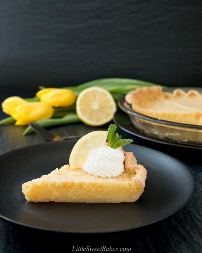 A slice of lemon pie on a black plate with whipped cream, lemon wedge and yellow tulips in the background.
