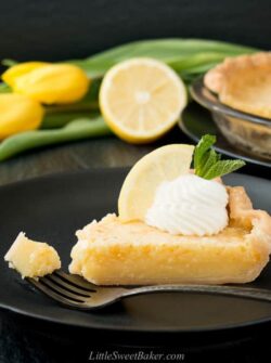 A slice of lemon pie on a black plate with piece on a fork.