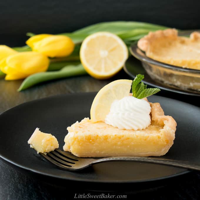 A slice of lemon pie on a black plate with piece on a fork.