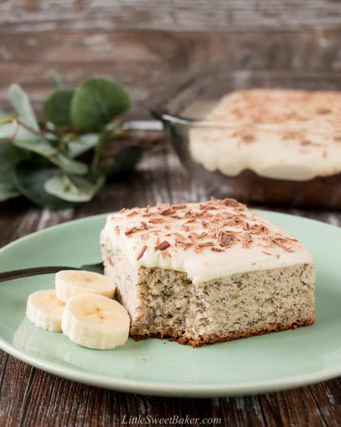 A slice of banana cake topped with cream cheese frosting on a green plate with some fresh banana slices.