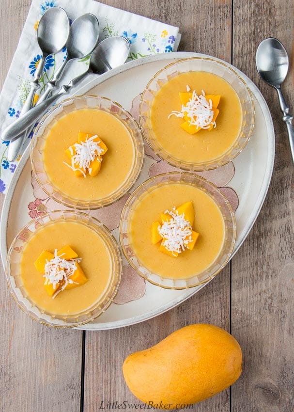 Four bowls of mango pudding on a plate surrounded by spoons and a mango.