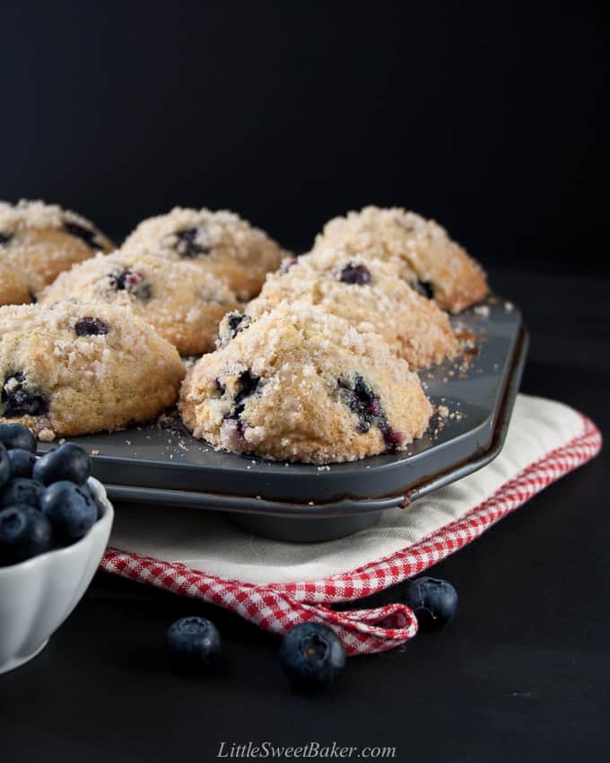 A muffin pan with blueberry muffins on a hot pad with a bowl of blueberries.