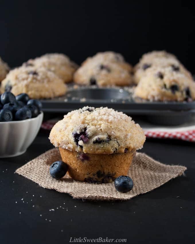 A blueberry muffin on a piece of burlap with a pan of muffins and blueberries in background.