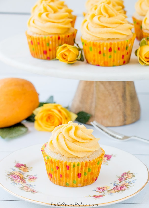 A mango cupcake on a white floral plate with a stand of cupcakes in the background.