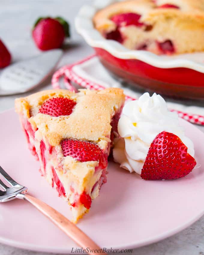 A slice of strawberry cake on a pink plate with some whipped cream and a strawberry half.