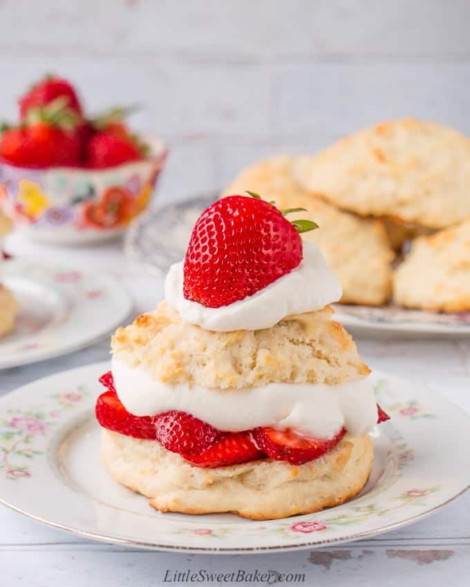 A strawberry shortcake on a vintage plate with biscuits and strawberries in the background.