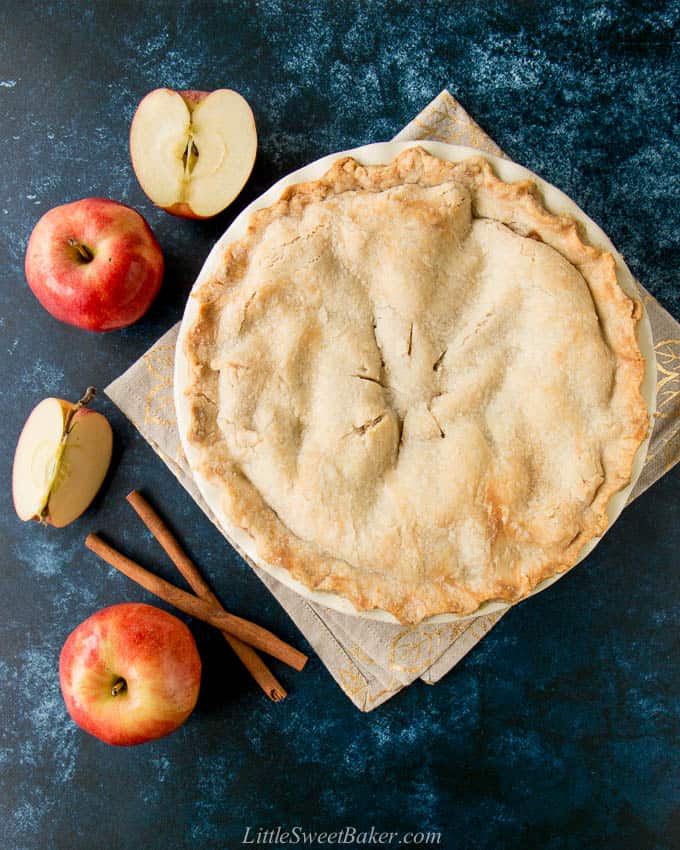 A whole apple pie on a beige napkin surrounded by apples and cinnamon sticks.