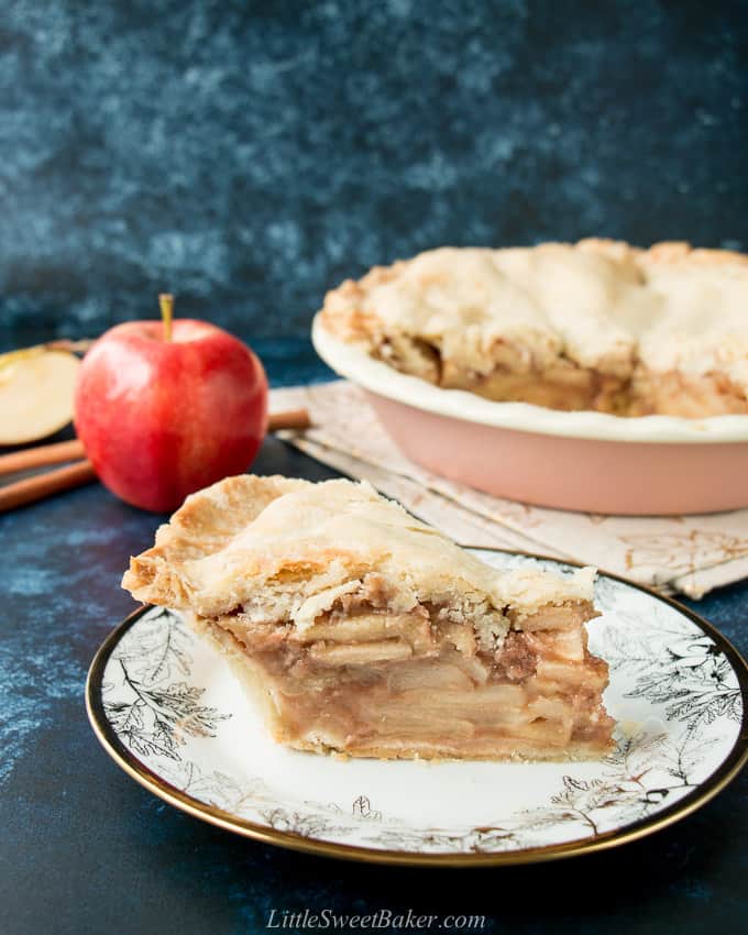 A thick slice on apple pie on a white and gold plate with the rest of the pie in the background.