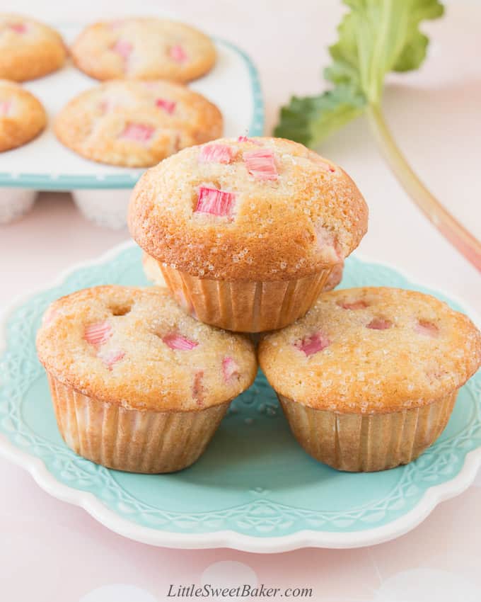 Three rhubarb muffins on a turquoise plate with a pan of muffins and rhubarb stalk in the background.