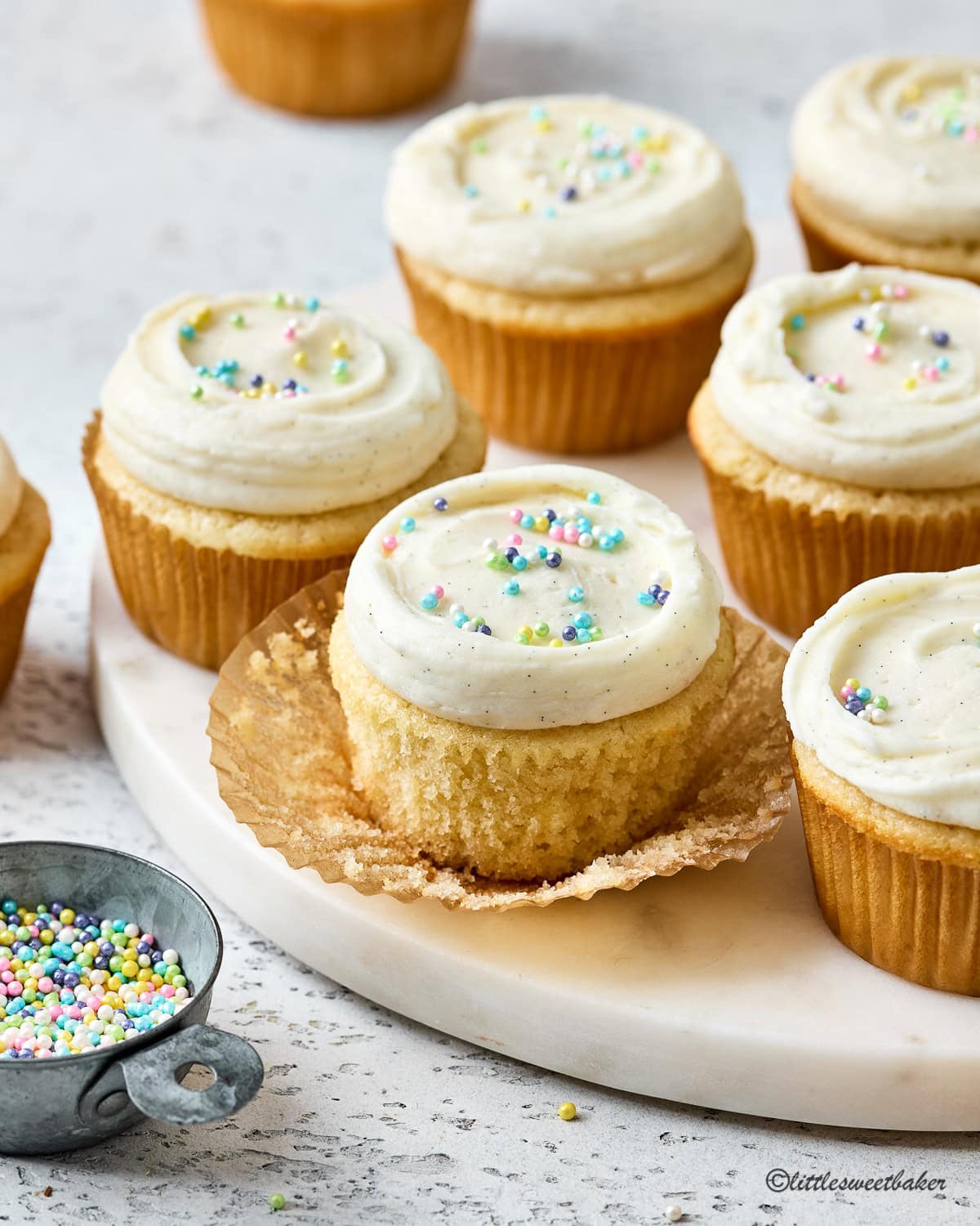 Vanilla cupcakes topped with vanilla buttercream and pastel sprinkles on a white marble.