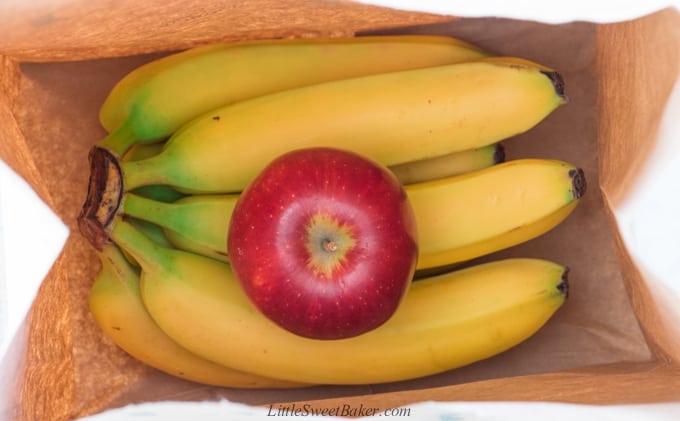 A bunch of bananas in a paper bag with a red apple.