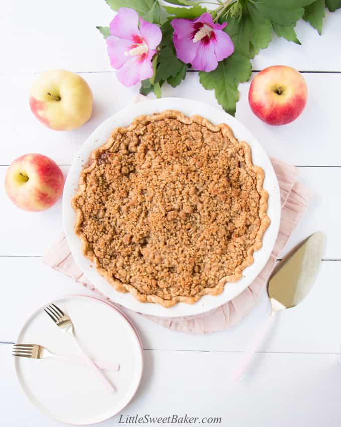 A Dutch apple pie on a pink napkin surrounded by flowers, apples, plate, and forks.