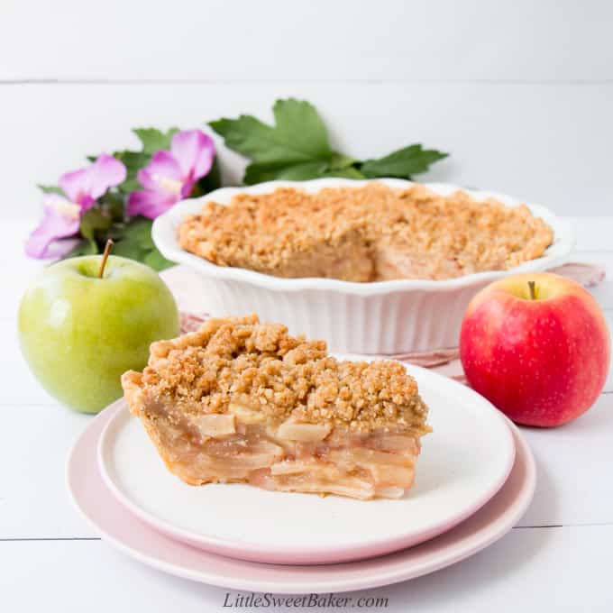 A slice of dutch apple pie on a white and pink plate with apples, the rest of the pie and flowers in the background.