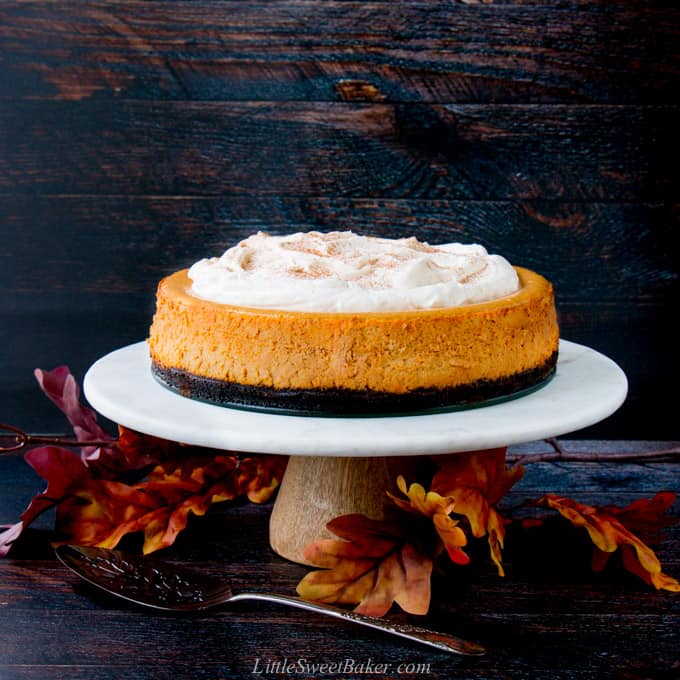 A pumpkin cheesecake on a marble cake stand with a dark wood background and fall leaves.