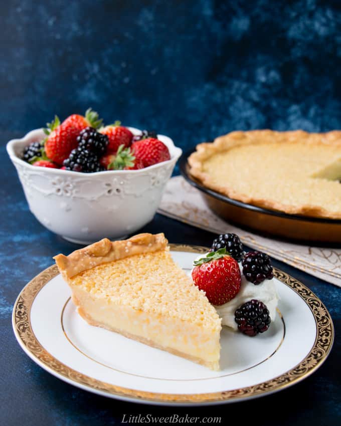 A slice of buttermilk pie on a white and gold plate with some berries and whipped cream.