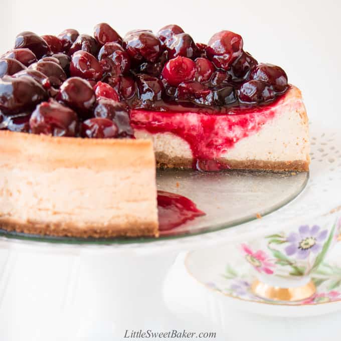 A cherry cheesecake on a white cake stand with a slice cut out.