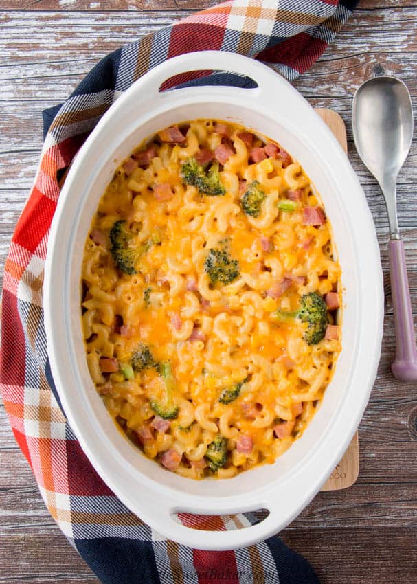 A leftover ham and vegetable casserole on a wooden board with plaid napkin and serving spoon.