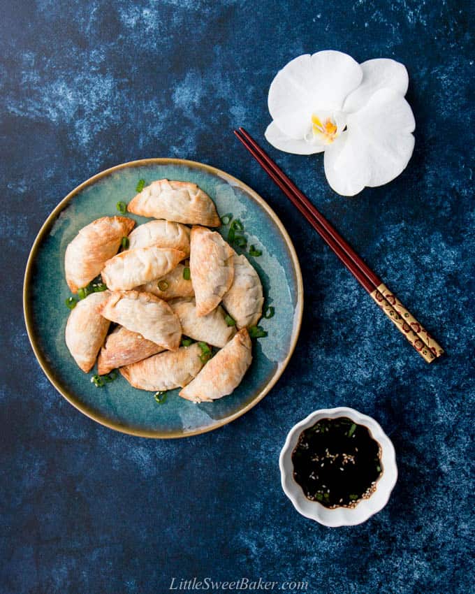 A plate of baked Chinese dumpling surrounded by a pair of chopsticks, soya sauce and a white orchid.