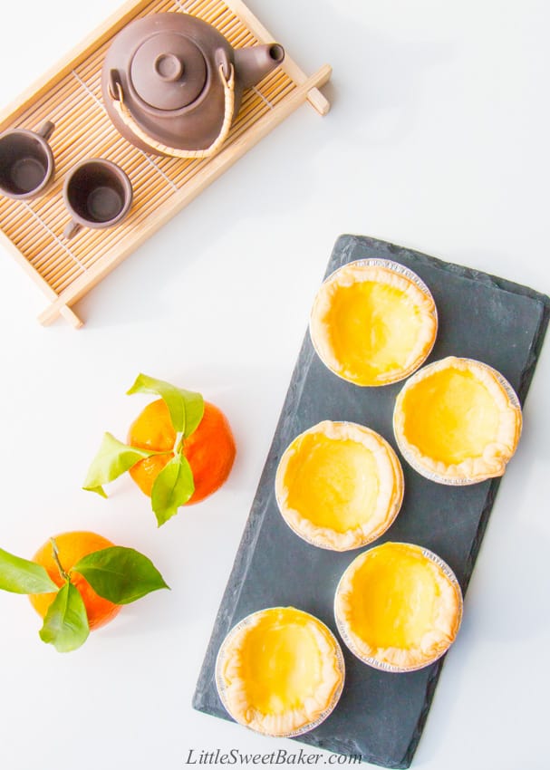 Chinese egg tarts on black late with mandarins and tea serving tray.