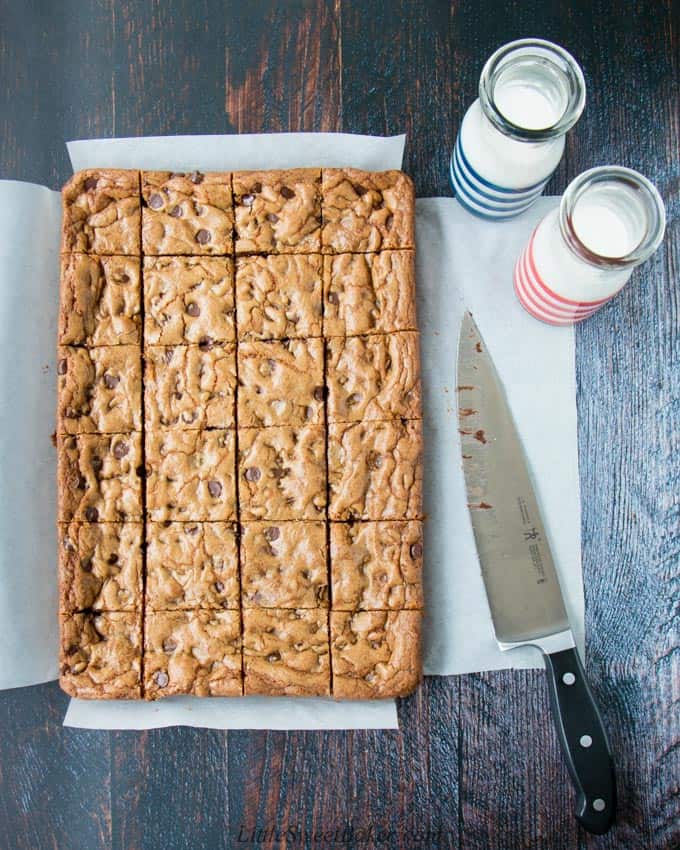 A tray of chocolate chip cookie bars cut into squares surrounded by a knife and two glasses of milk.