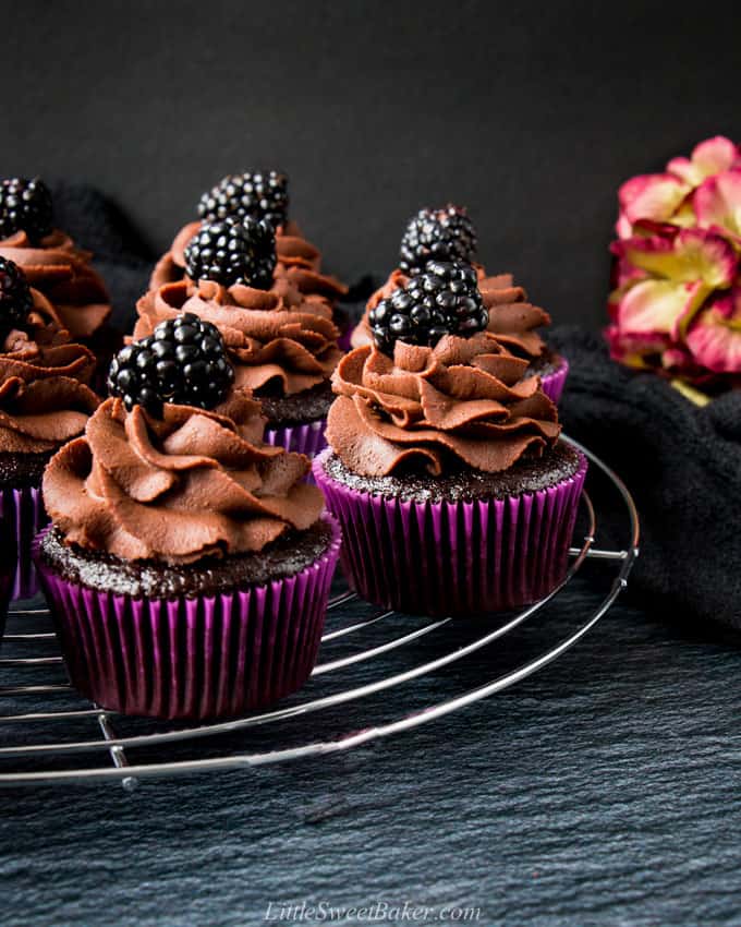 chocolate cupcakes in purple paper liners topped with chocolate ganache frostings and blackberries on a cooling rack.