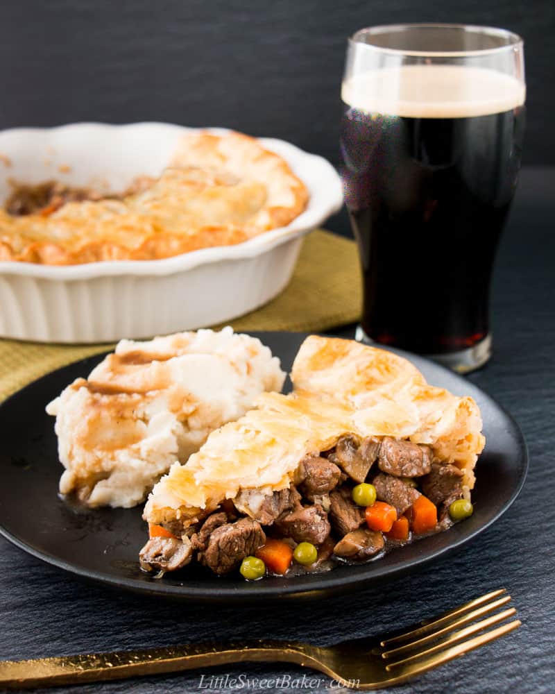 A slice of steak and stout pie with a side of mashed potatoes and gravy with a pint in the background.