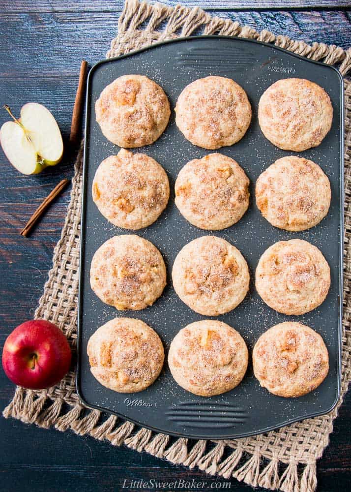 A muffin pan of apple cinnamon muffins on a burlap mat with apples apples and cinnamon sticks