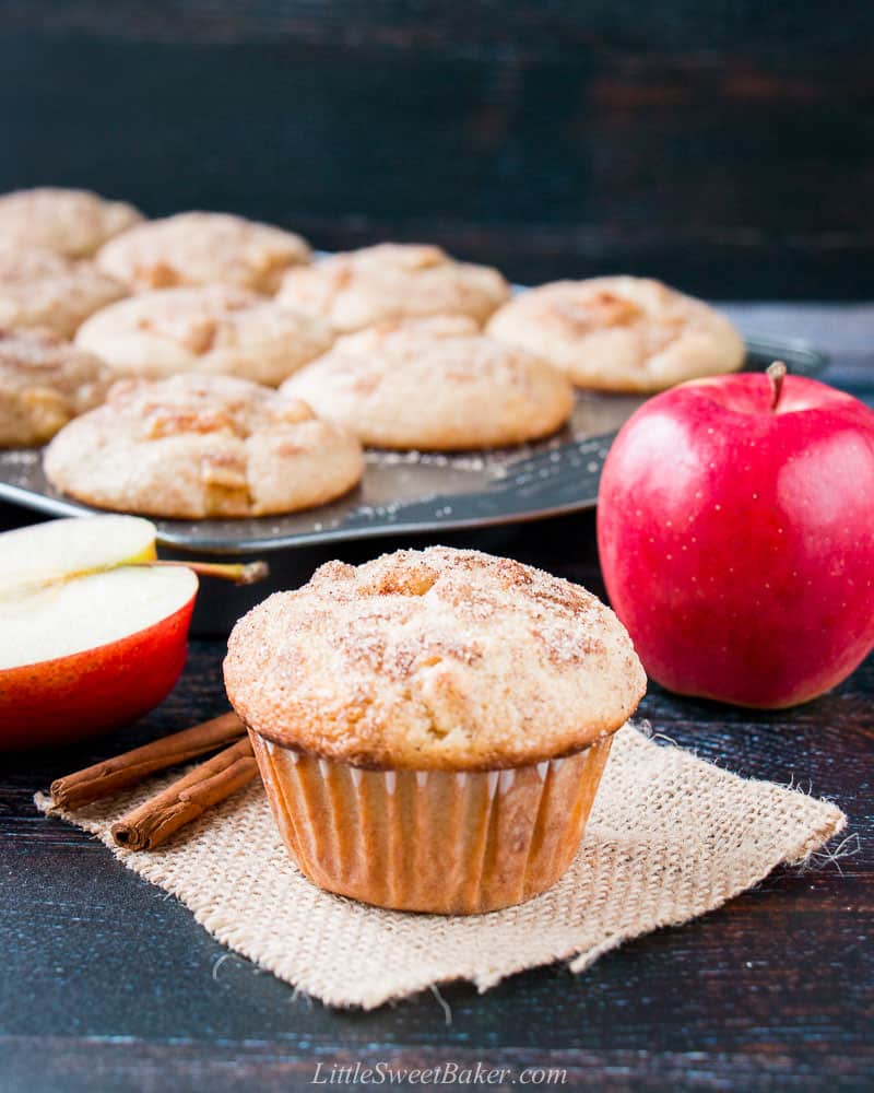 An apple cinnamon muffin in a piece of burlap with two cinnamon sticks, red apples and muffin pan in the background.