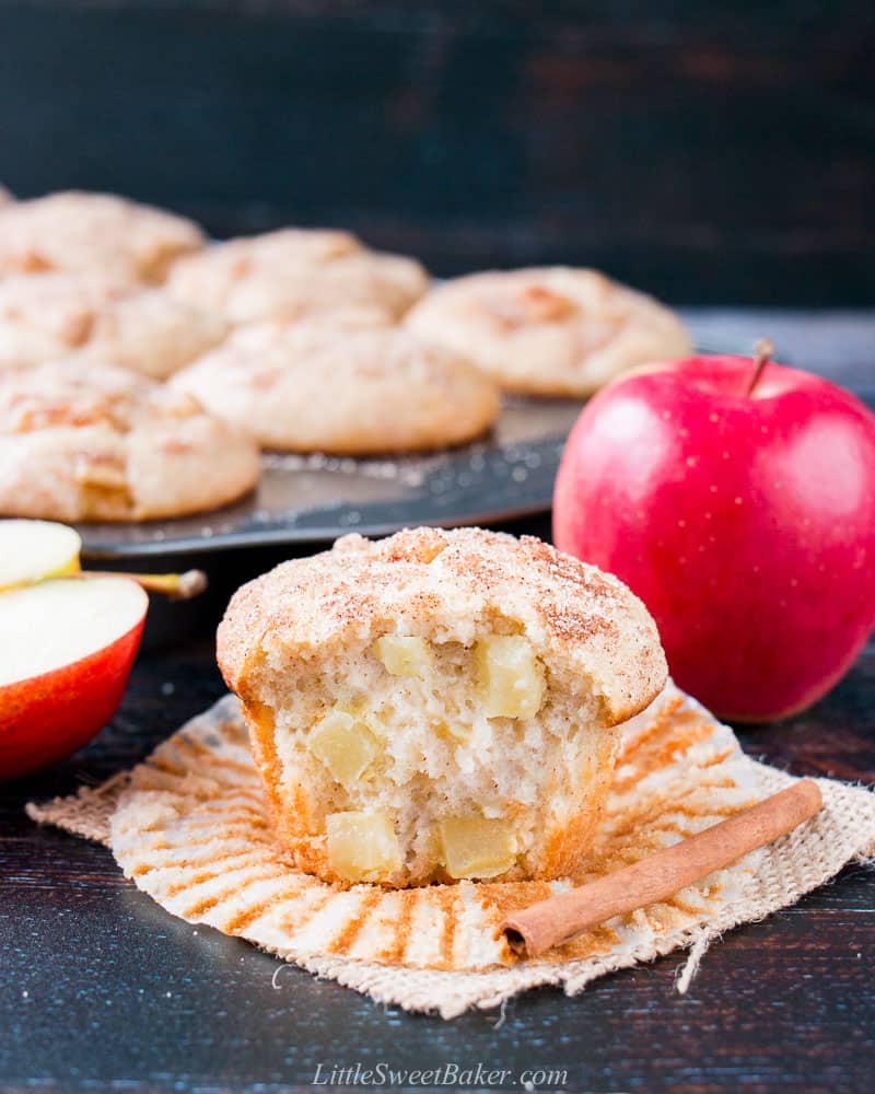 An apple muffin with a big bite taken out to show the inside texture with a cinnamon stick, apples, and muffin pan in the background.