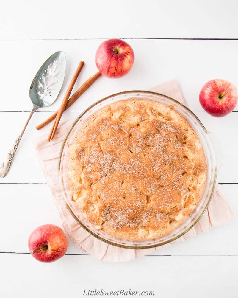A cinnamon sugar apple cake on a pink ruffled cloth surrounded by apples and cinnamon sticks.