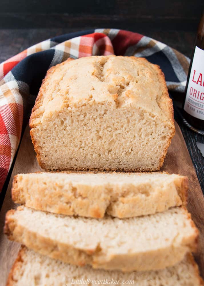 A loaf of beer bread on a wooden cutting board with a plaid kitchen towel and bottle of lager.