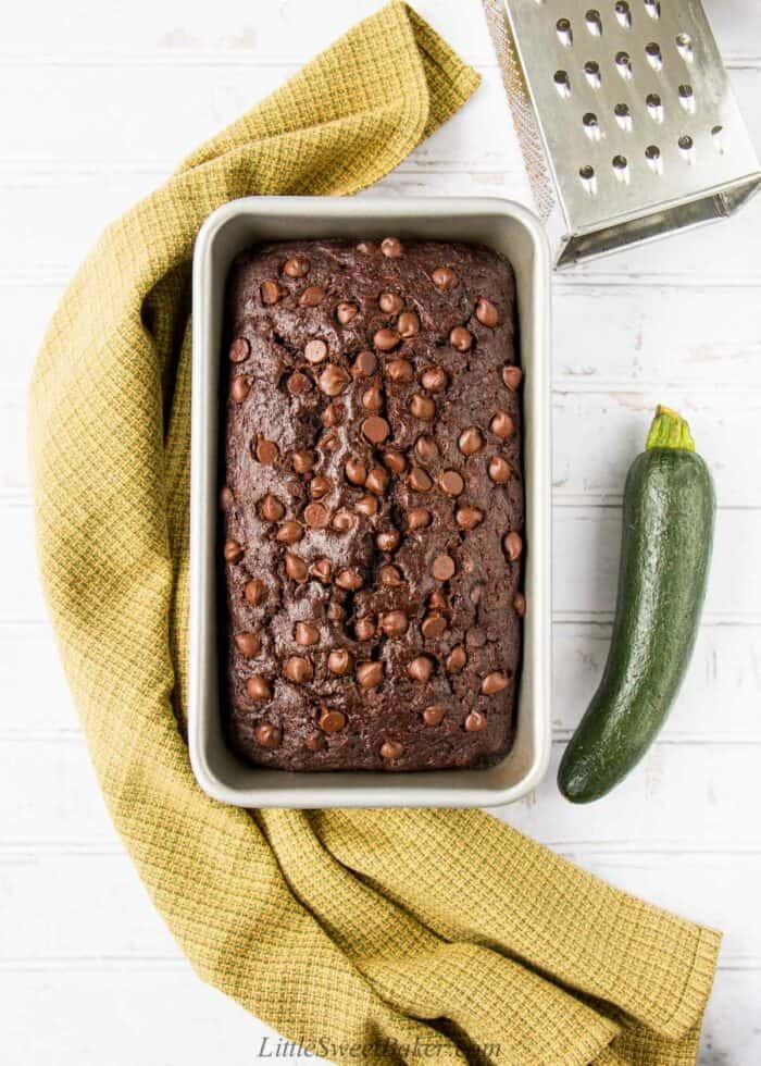 A loaf of chocolate zucchini bread with a green dish towel, zucchini and grater around it.