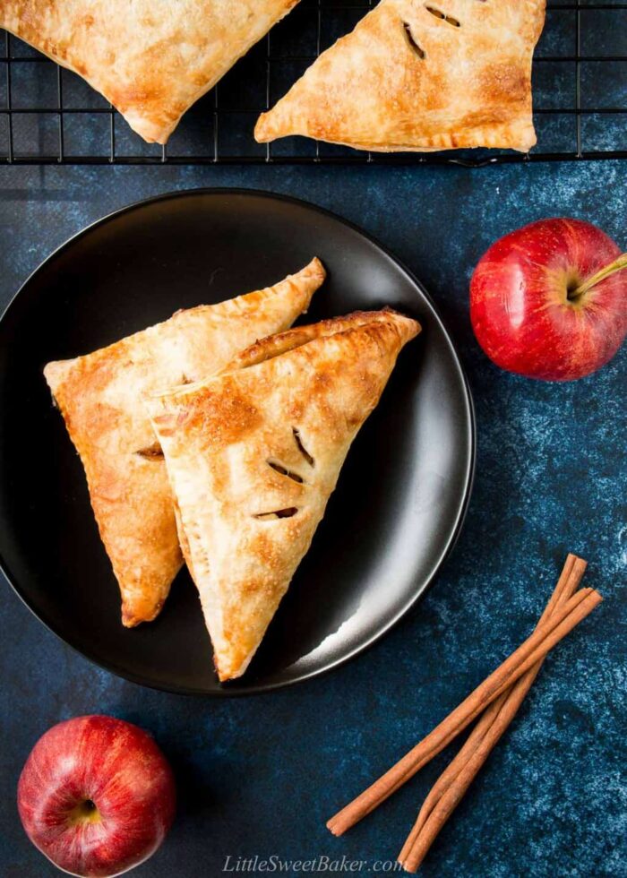 Puff pastry apple turnovers on a black plate with apple and cinnamon stick around.