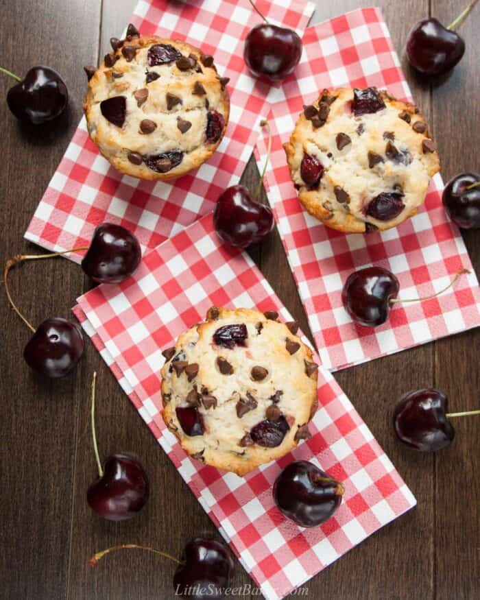 An overhead view of cherry chocolate chip muffins on red plaid napkins and surrounded by black cherries.