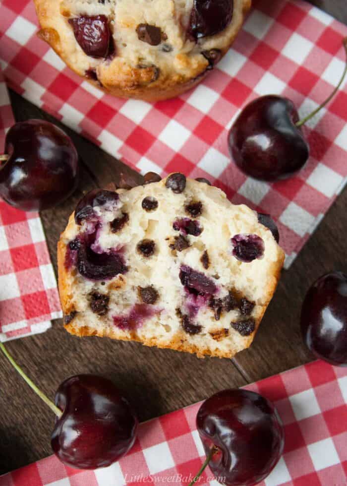 Half of a cherry chocolate chip muffin on a wooden table surrounded by cherries and red plaid napkins.