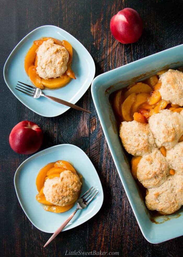 Homemade peach cobbler in a tray and two serving plates on a wooden table surrounded by two peaches.